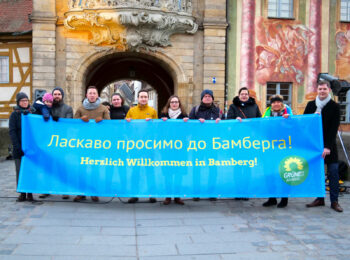 Grüne Mitglieder halten und stehen hinter blauem Banner mit der Aufschrift "Herzlich willkommen in Bamberg" auf Ukrainische und Deutsch