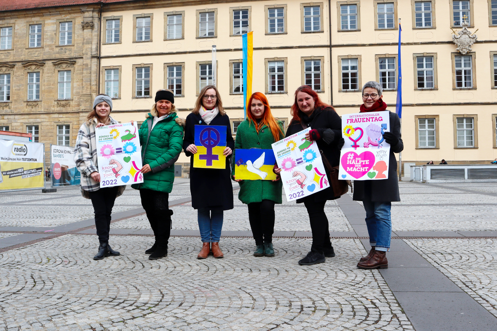 sechs grüne Frauen stehen nebeneinander auf dem Maxplatz in Bamberg und halten Schilder mit feministischen Botschaften in die Kamera