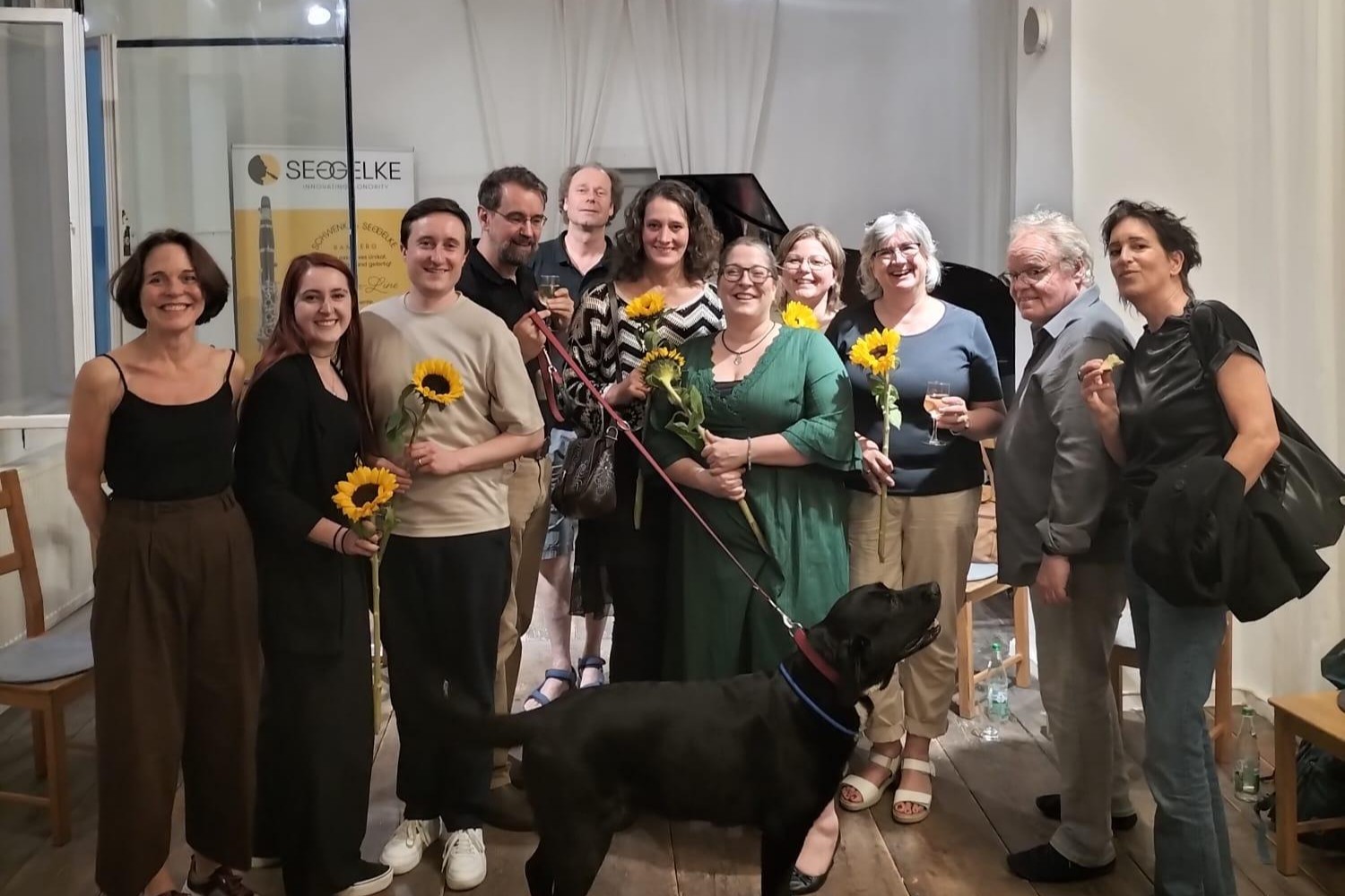 Gruppenbild der 11 Vortragenden vor einem schwarzen Piano. Im Vordergrund steht ein schwarzer Hund.