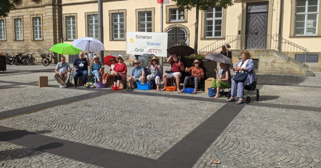 Mitglieder von Silbergrün sitzen vor dem Rathauseingang am Maxplatz mit Sonnenhüten, unter Sonnenschirmen und mit den Füßen in Wasserschüsseln.