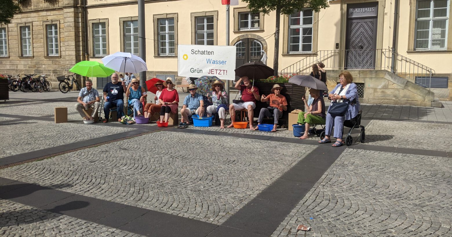 Mitglieder von Silbergrün sitzen vor dem Rathauseingang am Maxplatz mit Sonnenhüten, unter Sonnenschirmen und mit den Füßen in Wasserschüsseln.
