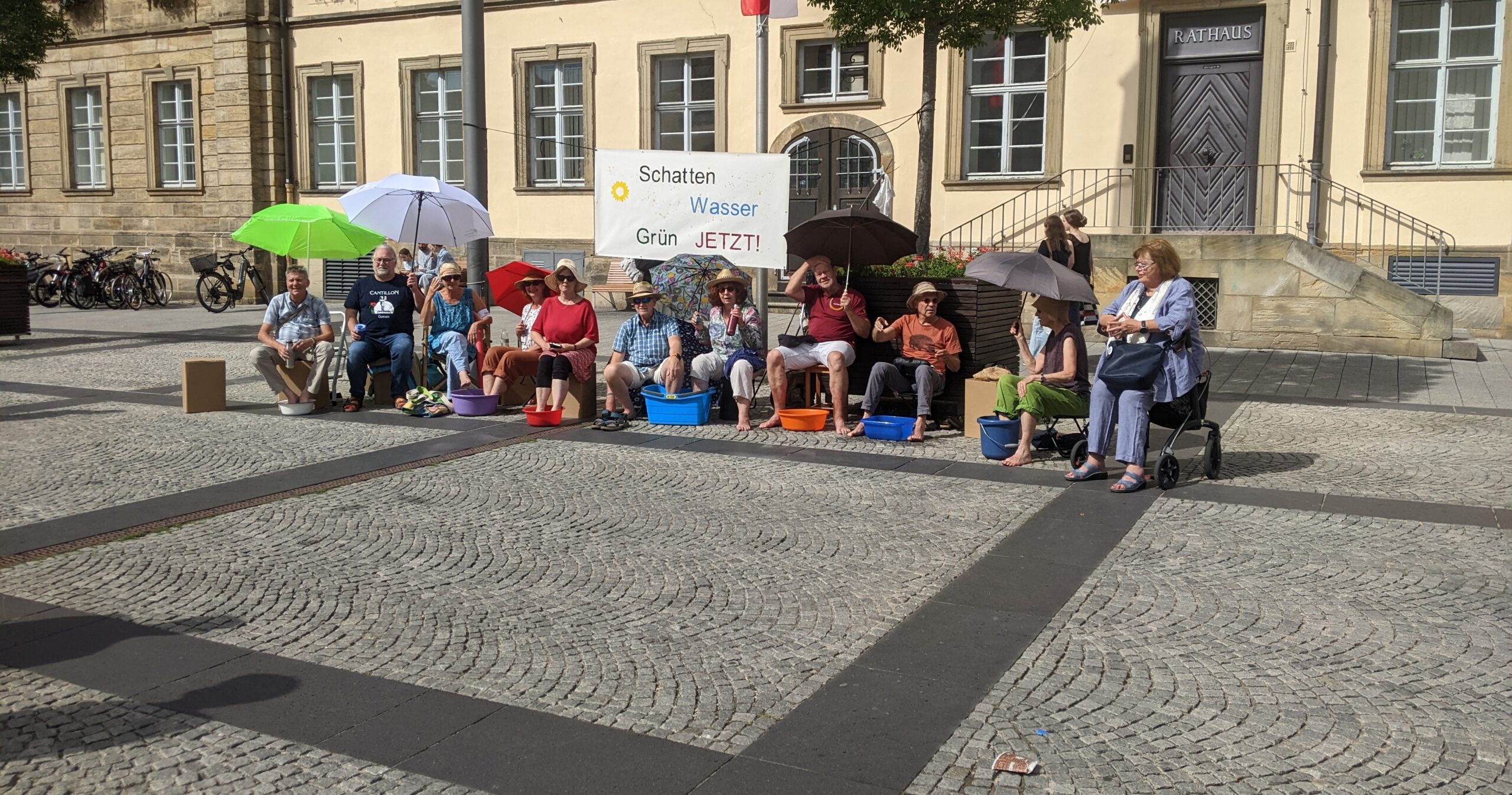 Mitglieder von Silbergrün sitzen vor dem Rathauseingang am Maxplatz mit Sonnenhüten, unter Sonnenschirmen und mit den Füßen in Wasserschüsseln.
