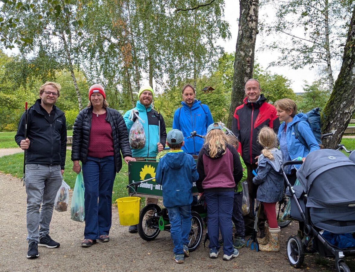 Eine Gruppe von sieben Erwachsenen und drei Kindern steht gemeinsam auf einem Kiesweg unter großen Birken. Die Personen tragen herbstliche Outdoor-Kleidung in gedeckten Farben sowie Blau und Rot. In der Mitte der Gruppe steht ein grüner Lastenfahrrad-Anhänger, der mit einem gelben Sonnenblumen-Logo und dem Schriftzug „Bamberg – mit...“ versehen ist. Neben dem Anhänger steht ein gelber Eimer, und einige Personen halten Müllbeutel in den Händen, was auf eine gemeinsame Aufräumaktion in der Natur hindeutet. Rechts im Bild ist zudem ein grauer Kinderwagen zu sehen.